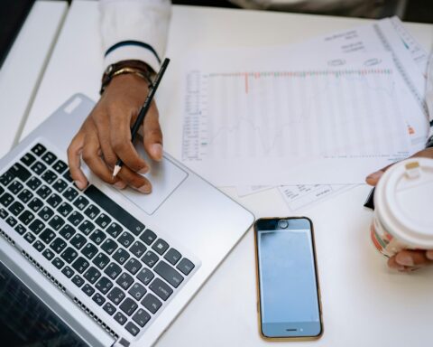 A businessman holding coffee and using a laptop at a desk with charts and a smartphone.